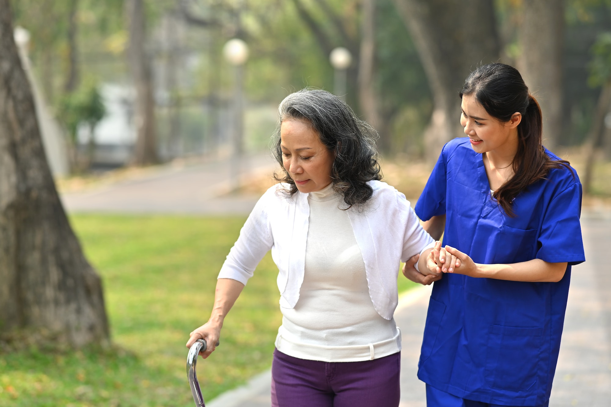 Young caring female nurse helping senior woman walking in the park.